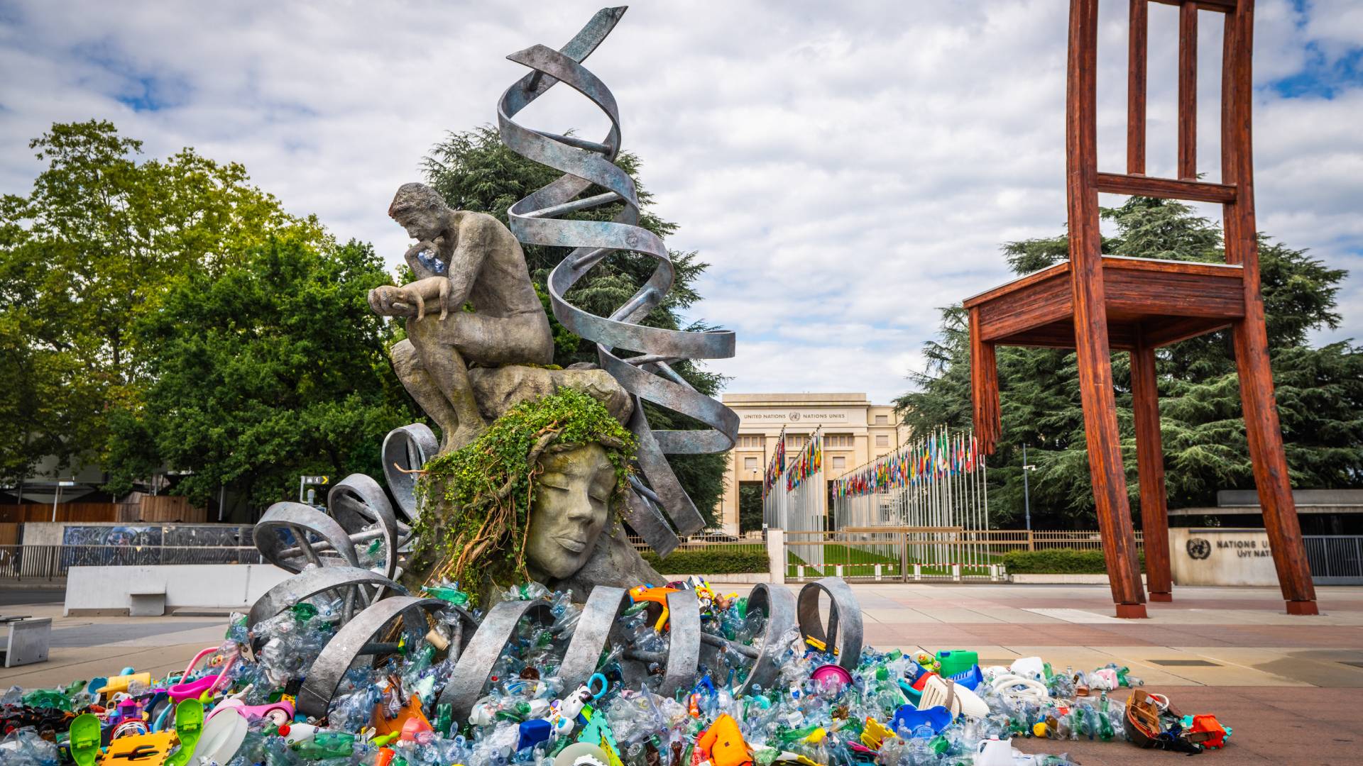 Male statue with his head in his hands sitting on a woman’s head, surrounded by metal coils, fresh flowers and waste plastic