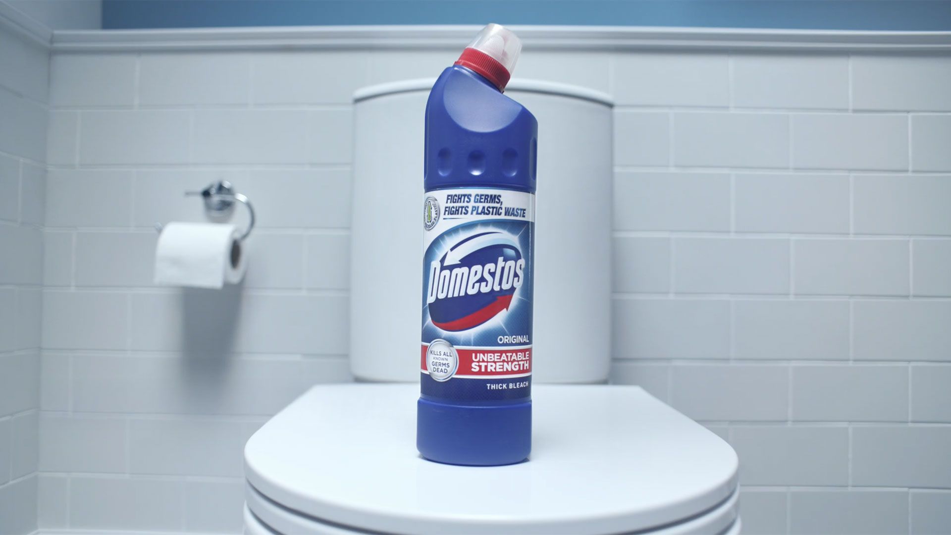 A blue bottle of Domestos Original thick bleach is positioned on the closed lid of a white toilet in a bathroom. The bottle has a red angled nozzle and a large red-and-blue branded label. The background features white tiled walls and a toilet paper holder.