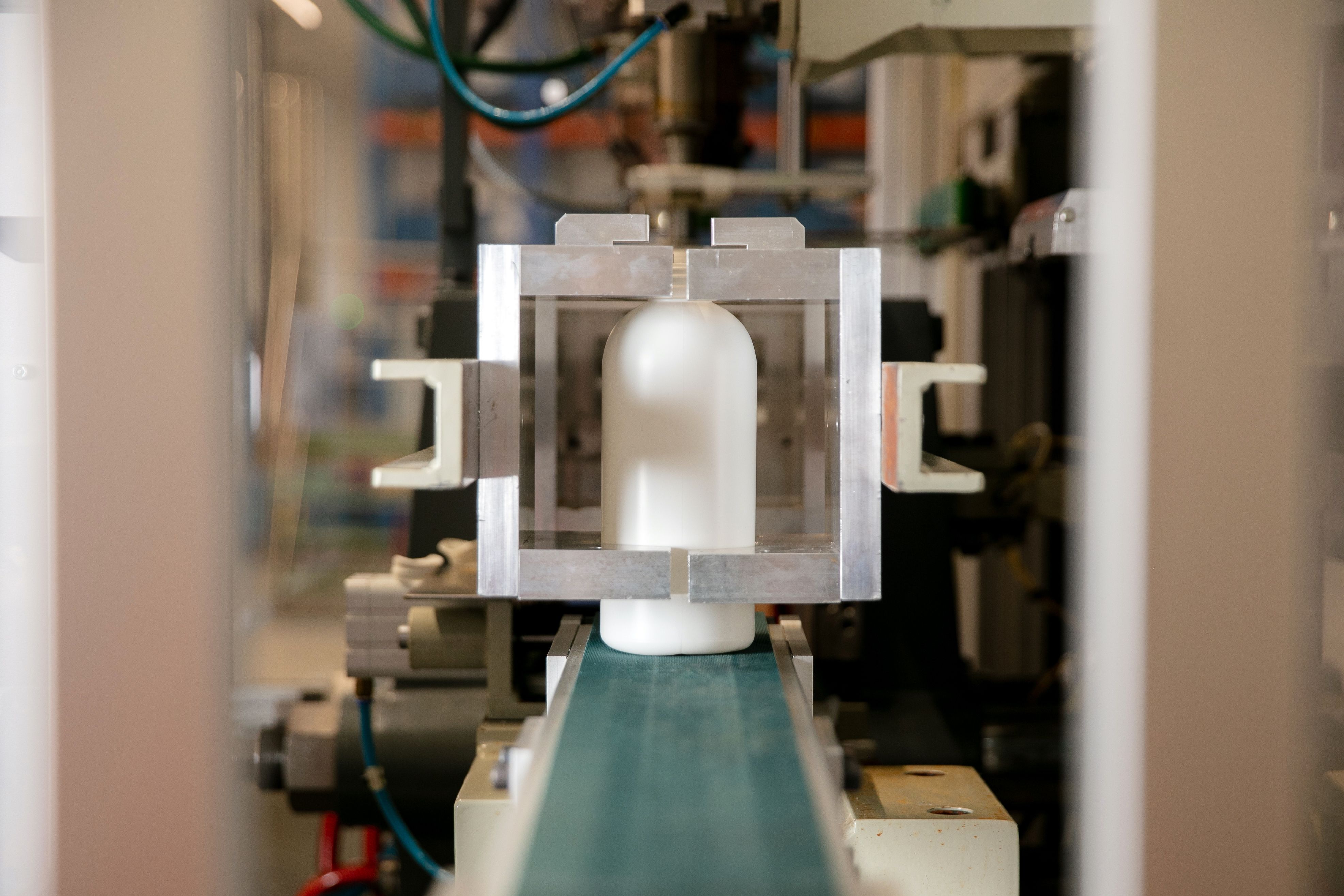 A close–up of a white plastic bottle in an industrial testing setting. The bottle is held in place by a rectangular metallic frame.