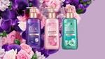 Three Lux perfumed hand soap bottles in purple, pink, and teal displayed against a background of vibrant roses and orchids on a pastel purple backdrop.