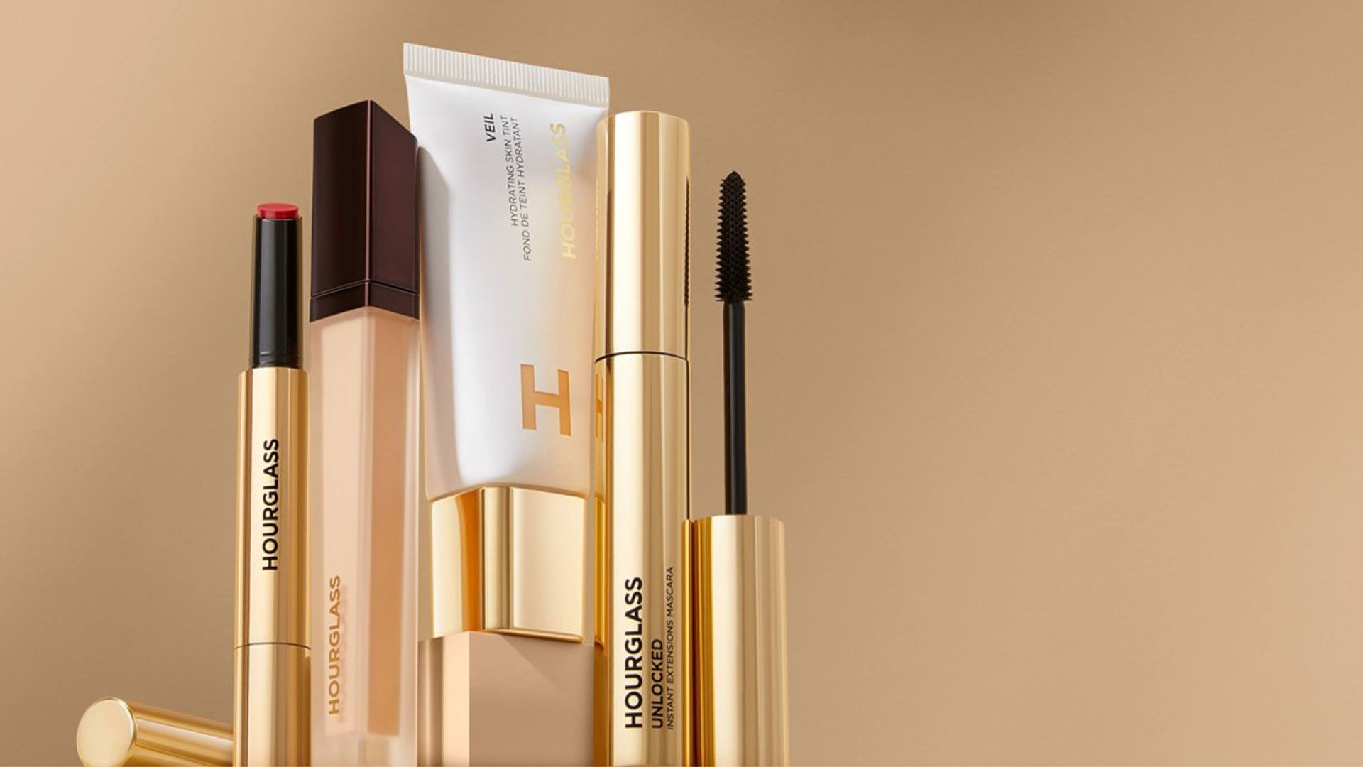 A display of Hourglass make-up products — including a lipstick, foundation, primer, and mascara — displayed standing upright against a warm beige background in gold and neutral-toned packaging.