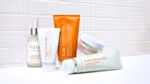 A selection of Kate Somerville skincare products arranged on a white surface against a tiled background. Items include a pump bottle of Liquid ExfoliKate, a white tube of ExfoliKate Intensive Exfoliating Treatment, an orange tube of ExfoliKate Glow Moisturiser, a white jar of Exfolikate Glow, and a green-and-white tube of ExfoliKate Cleanser.