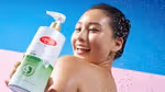 A person stands under splashing water against a bright blue background while holding a large bottle of Lifebuoy Anti Acne body wash in the foreground. The bottle has white and green packaging and features aloe vera imagery.