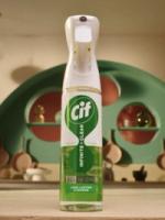 Bottle of Cif Infinite Clean all-in-one cleaning spray with green liquid, placed on a countertop with a decorative green wall and kitchen utensils in the background.