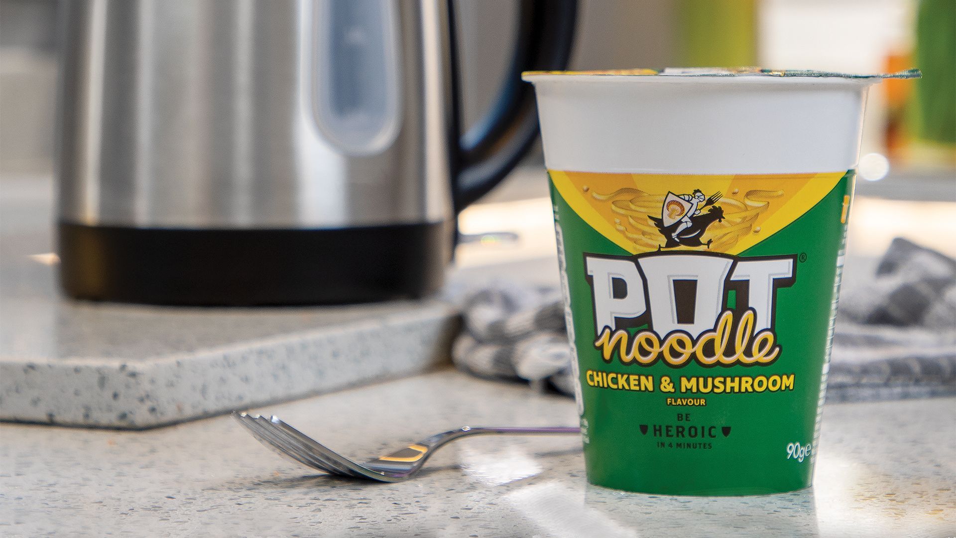 A Chicken & Mushroom Pot Noodle in a paper pot stands on a kitchen counter next to a kettle and a fork. 