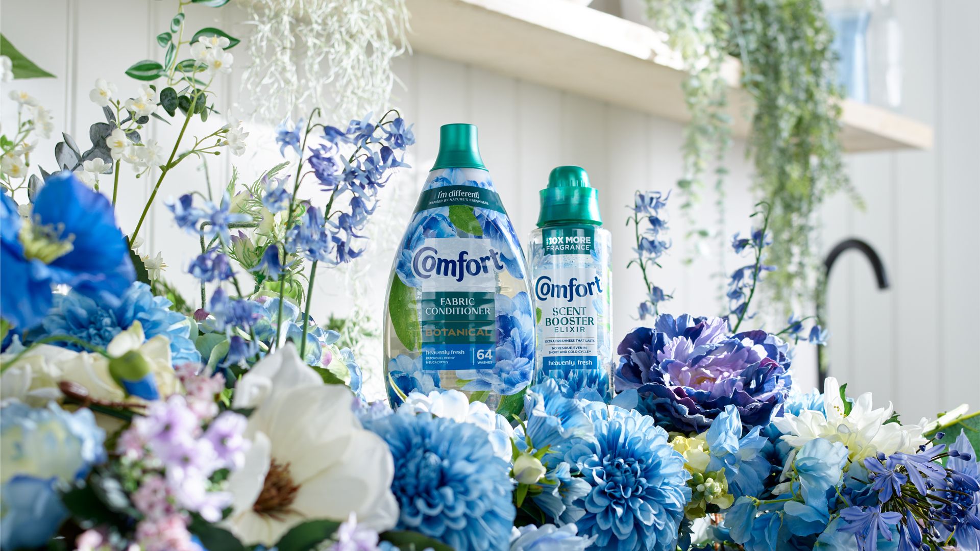 A bottle of Comfort Botanicals and Comfort Elixir – both in blue packaging – on a work surface surrounded by blue and white flowers.