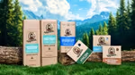 Dr. Squatch grooming products – including shampoo, conditioner, deodorant, and soaps – arranged on logs and grass in a natural outdoor setting with trees, mountains, and a blue sky in the background.