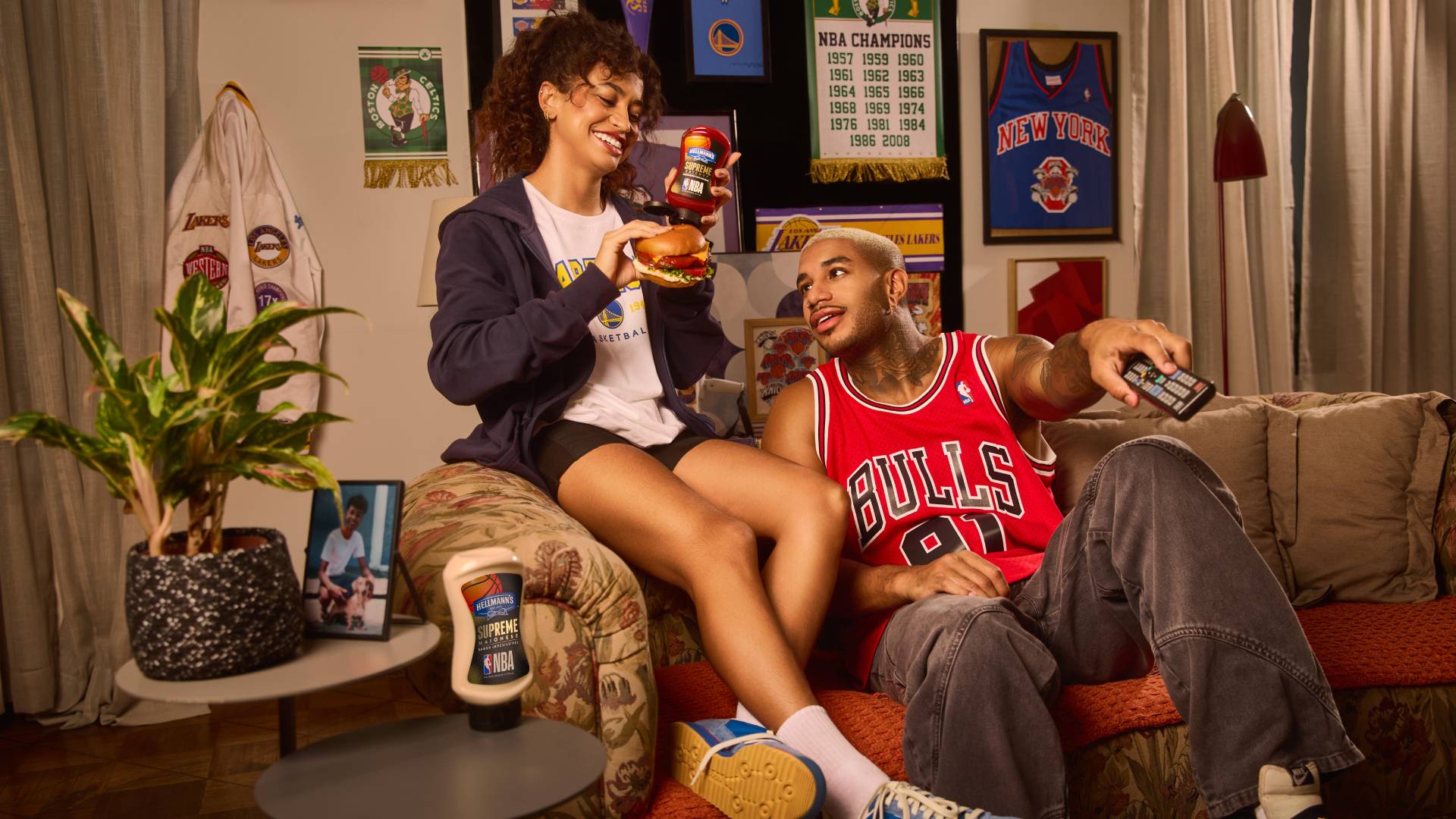A young man in a Red Bull’s NBA team shirt smiles at a young woman as she squeezes a Hellmann’s flavoured mayo on a burger