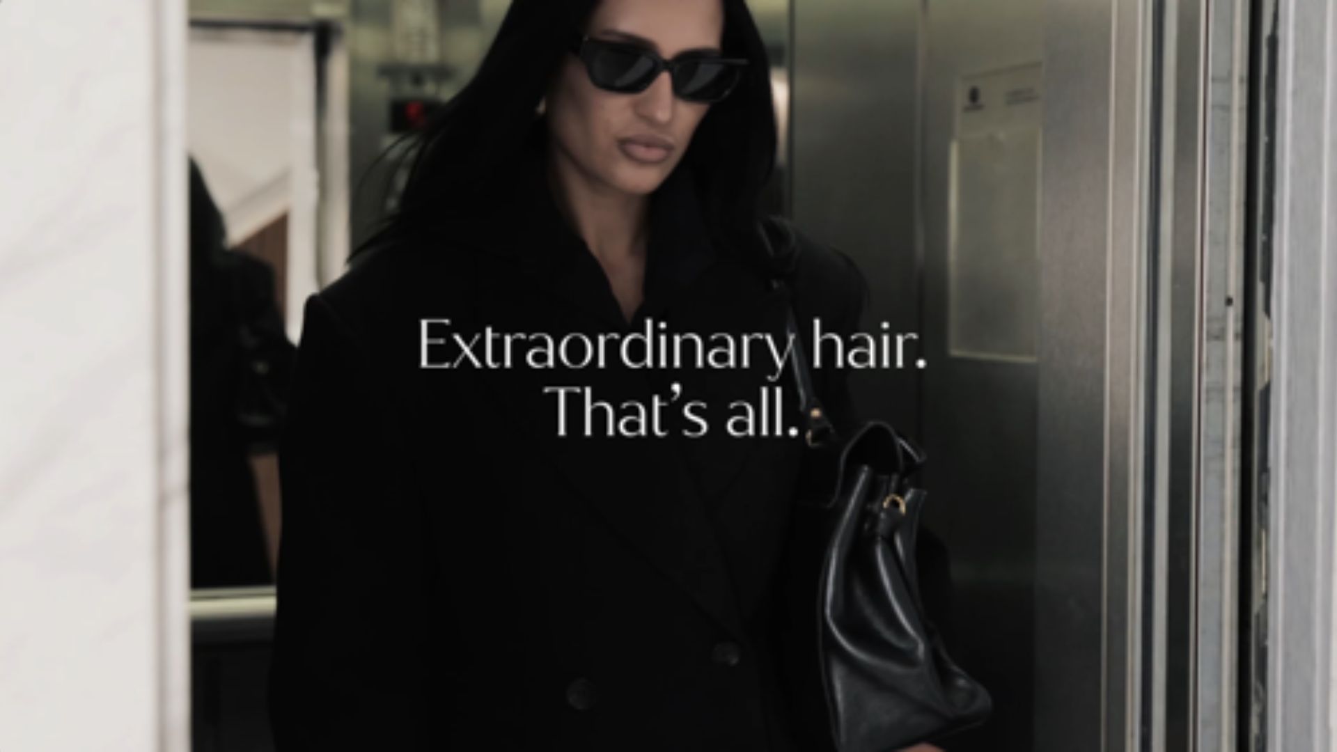 A person wearing a dark coat and sunglasses stands indoors holding a black handbag, with long dark hair visible. Overlaid text reads: “Extraordinary hair. That’s all.”