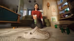 A person is kneeling on a carpeted floor, holding a can of Cif Infinite Clean aimed at a dirty white shoe in the foreground. The room has a cozy decor with a green couch, wooden furniture, and various items scattered around.