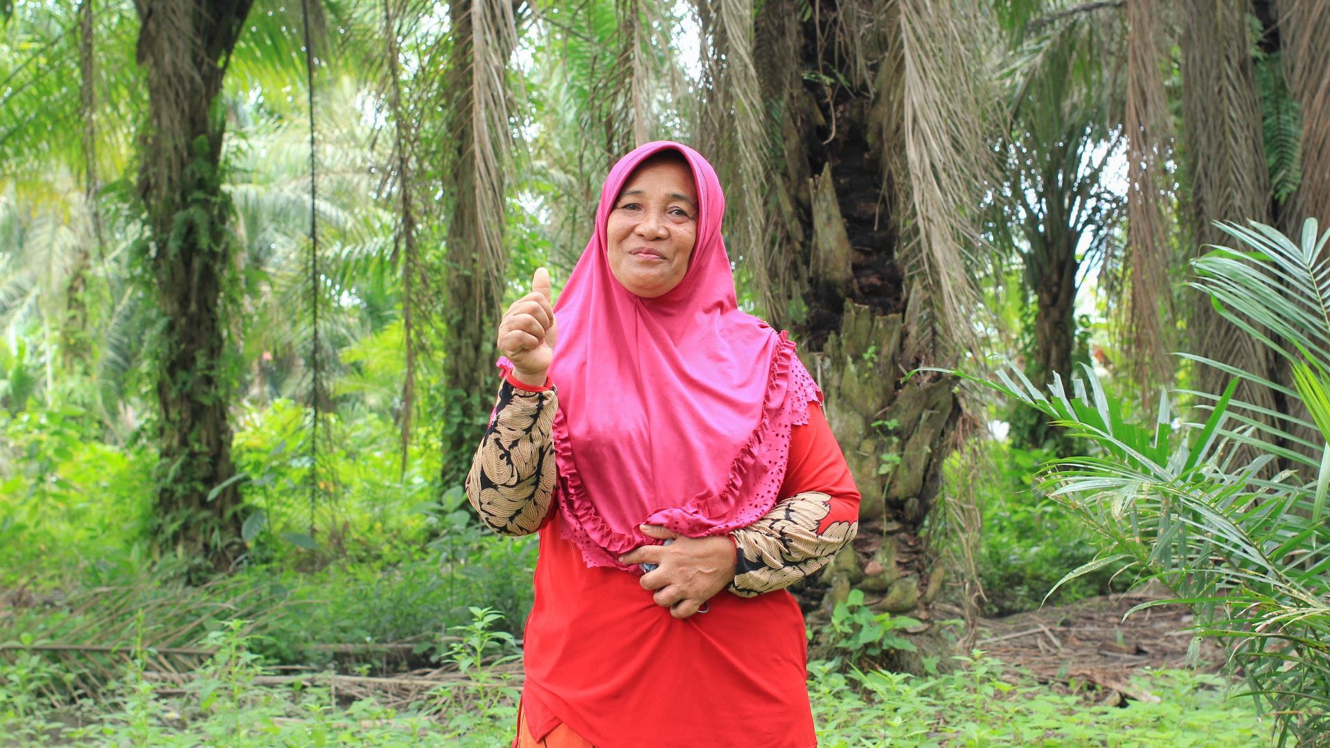 A smiling woman standing in a palm plantation, wearing a bright pink headscarf and a red outfit with patterned sleeves. She’s giving a thumbs-up gesture, surrounded by vibrant greenery and palm trees.