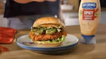 Close-up of a crispy fried chicken sandwich on a plate, layered with lettuce, pickles, and avocado slices inside a soft bun. A bottle of Hellmann’s Spicy Mayonnaise Dressing is placed to the right, and a red napkin is partially visible on the left. The setting appears to be a kitchen countertop with warm lighting.