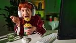 A person wearing large white headphones is eating instant noodles from a cup labelled 'Knorr' while sitting at a desk. The desk has a glass of water, a white computer keyboard, and a mouse and we can see the back of a computer monitor. The background includes shelves with books and plants, and the scene is lit with green ambient lighting.