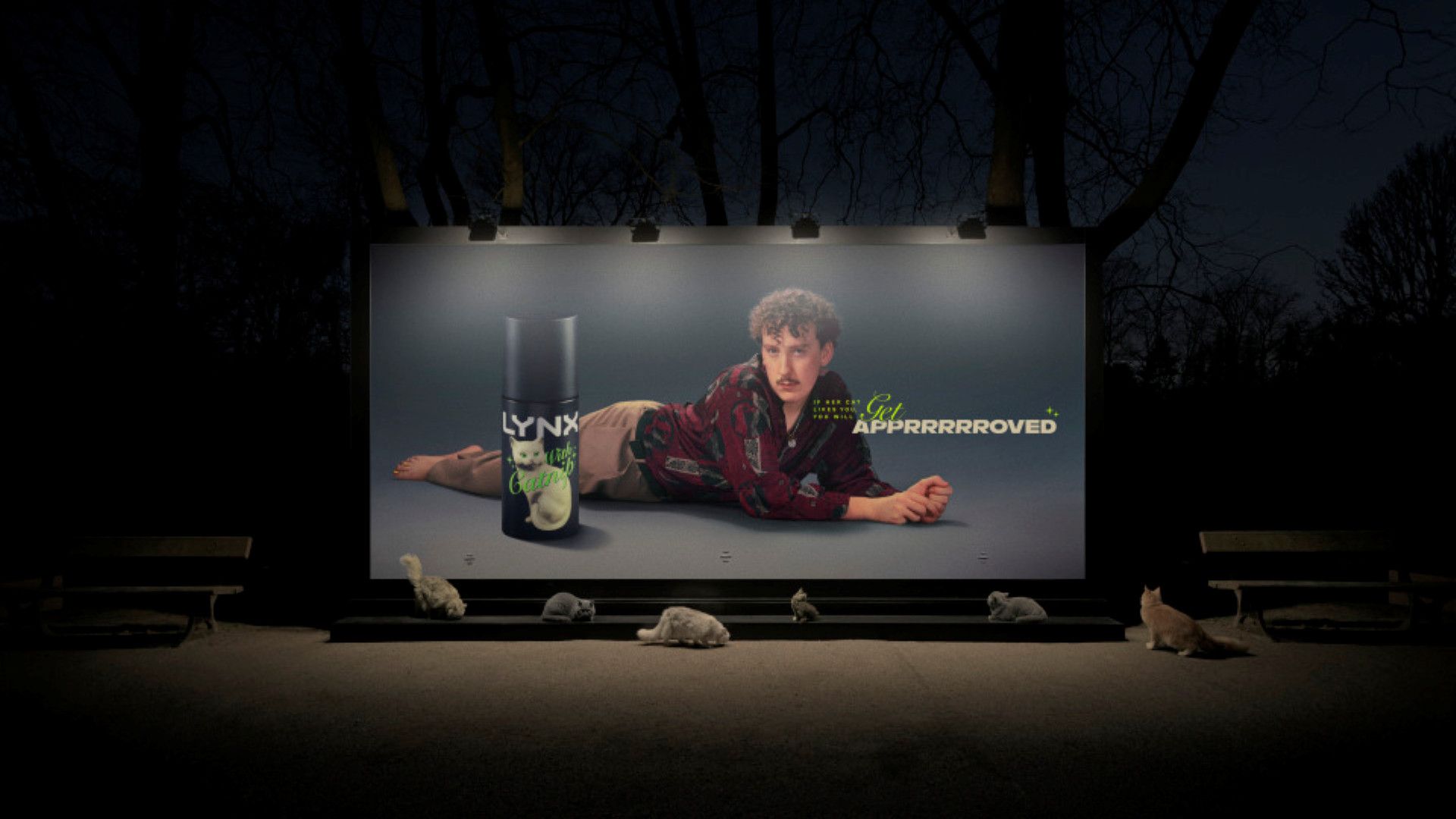 A billboard features an advert of a man with a moustache and curly hair lying on the floor behind a can of Axe Catnip. The image says: If her cat likes you, you will get apprrrroved. The billboard is scented with Axe Catnip and below it you can see six cats.