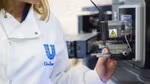 A close-up photo of a scientist in a Unilever-branded white lab coat, looking at a large digital microscope.