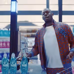 Usain Bolt standing at a checkout counter in a brightly lit store, wearing a patterned jacket and white shirt, holding a bottle of Persil detergent. Shelves in the background are stocked with colourful bottles of cleaning products in pink, blue, and turquoise.