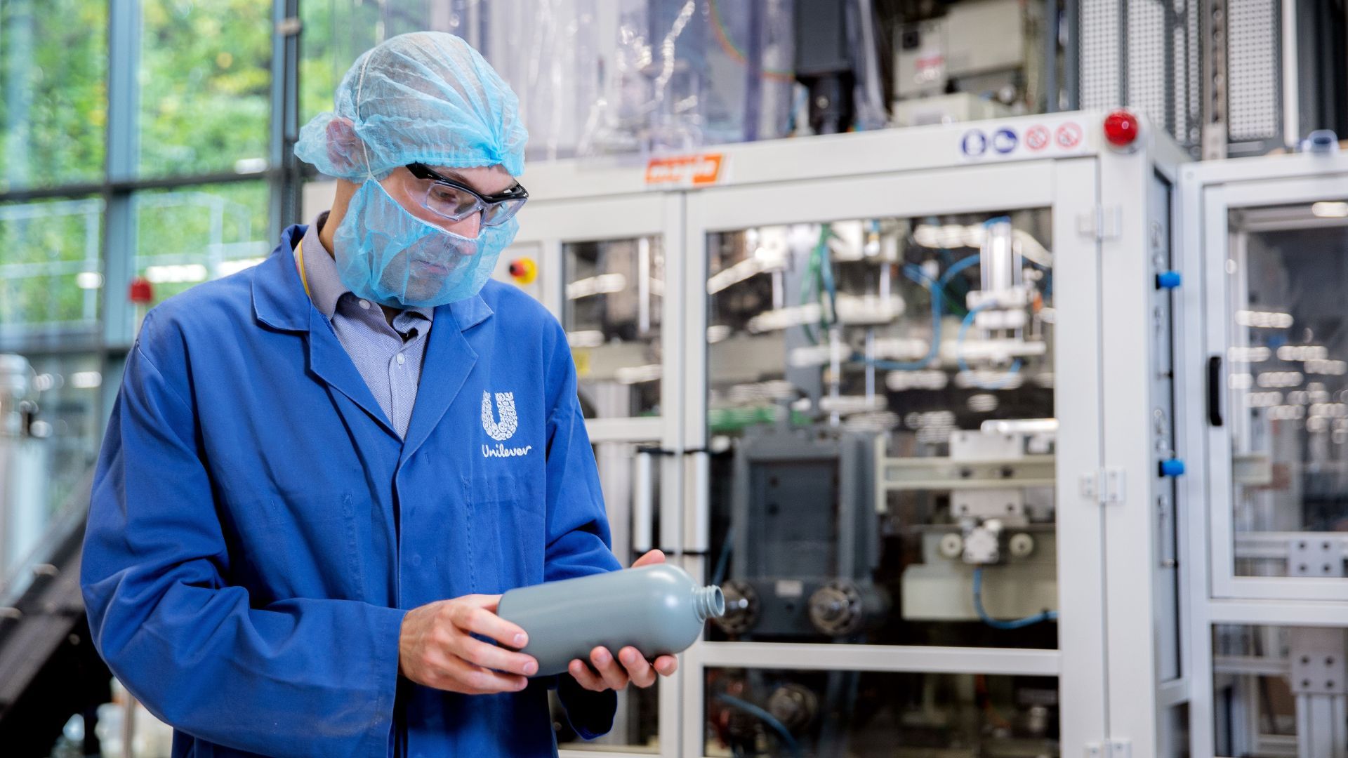 A Unilever packaging expert wearing protective clothing and glasses in an industrial setting, inspecting a plastic bottle  in front of manufacturing or packaging machinery. The Unilever logo is visible on the person’s coat.
