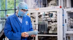 A Unilever packaging expert wearing protective clothing and glasses in an industrial setting, inspecting a plastic bottle in front of manufacturing or packaging machinery. The Unilever logo is visible on the person’s coat.
