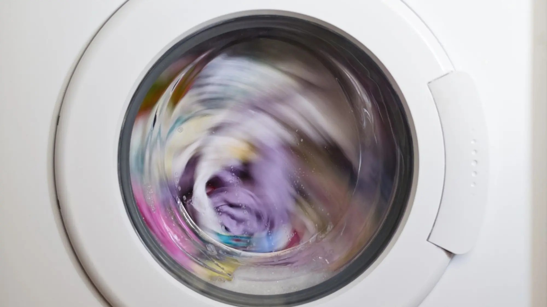 Zoomed in shot of a white washing machine with colourful clothes inside during a wash cycle.