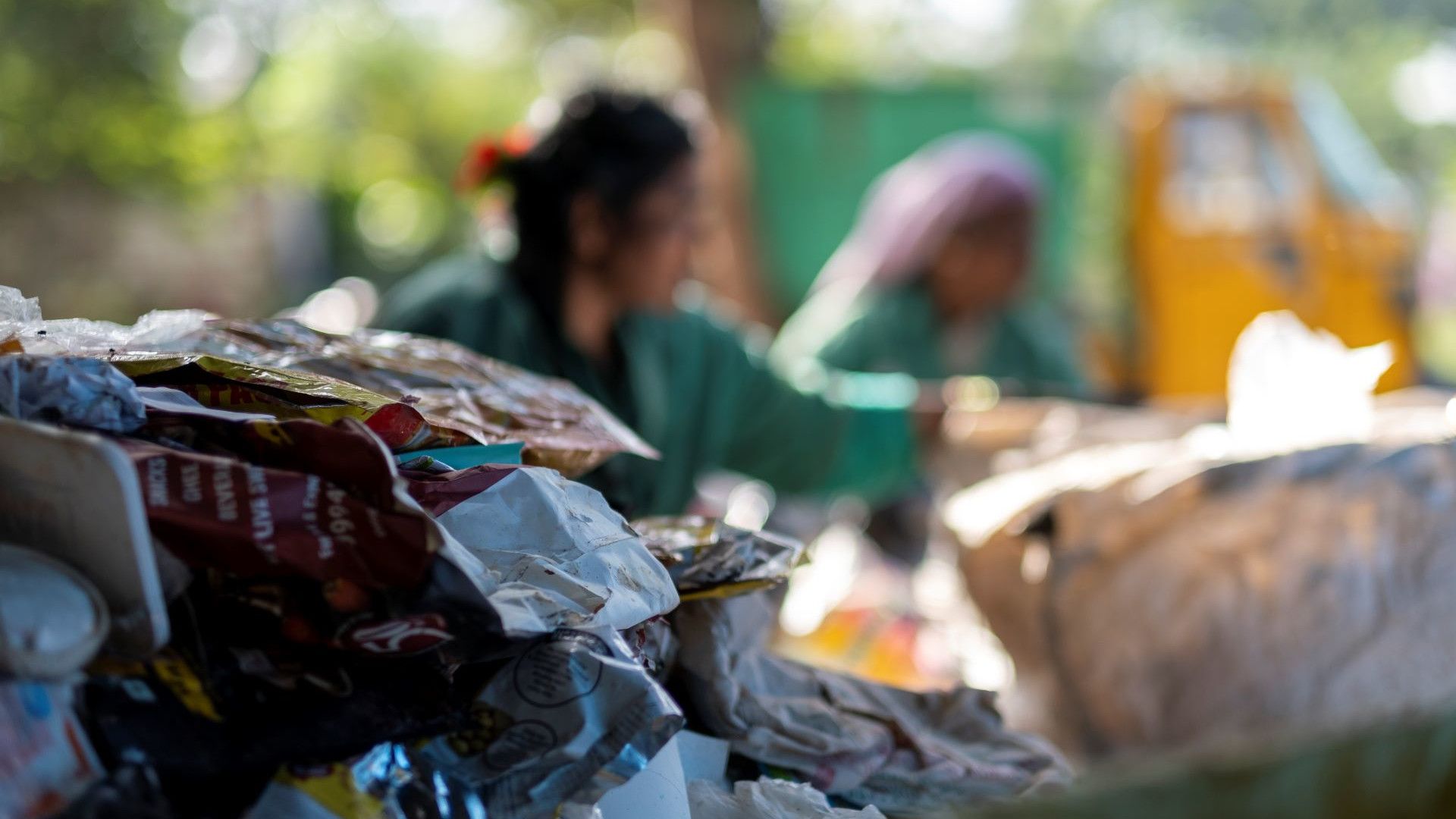 A close-up of a pile of discarded plastic packaging, with two people sorting waste in the blurred background.