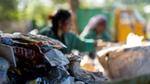 A close-up of a pile of discarded plastic packaging, with two people sorting waste in the blurred background.