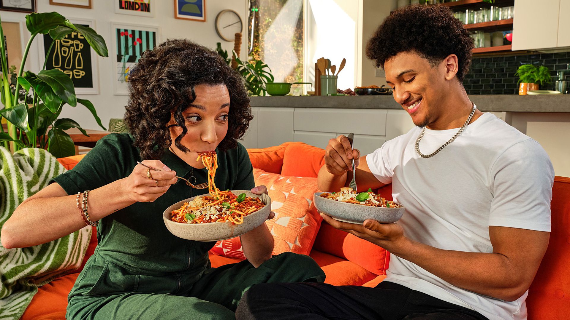 Two people sitting on an orange couch in a cozy living room, enjoying bowls of pasta together. The room is decorated with plants and artwork.
