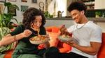 Two people sitting on an orange couch in a cozy living room, enjoying bowls of pasta together. The room is decorated with plants and artwork.
