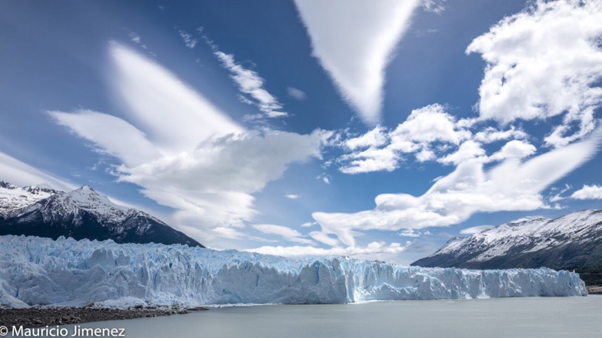 View of the terminus of a glacier as seen from sea level with mountains in the background and a bright, sunny sky