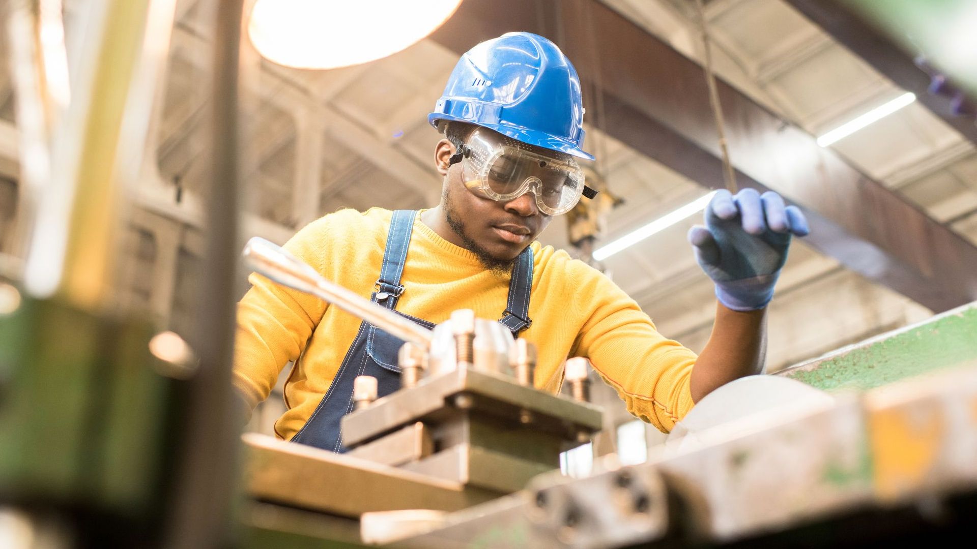 A man wearing a blue hard-hat, protective glasses and gloves working in a factory.