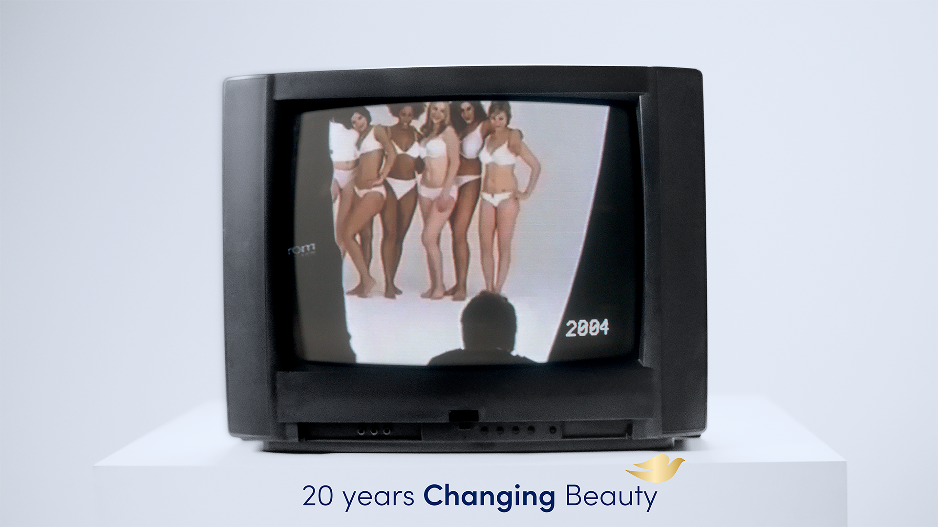 A TV resting on a white plinth, showing a 2004 ad from Dove. The ad on the screen shows six women in their underwear. The text at the bottom of the image reads: ‘20 years Changing Beauty’.