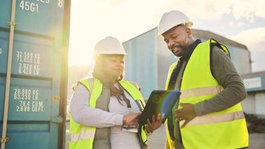 Two workers using a digital tablet discussing ecommerce, logistics, supply chain and shipping. Freight and cargo in the background.