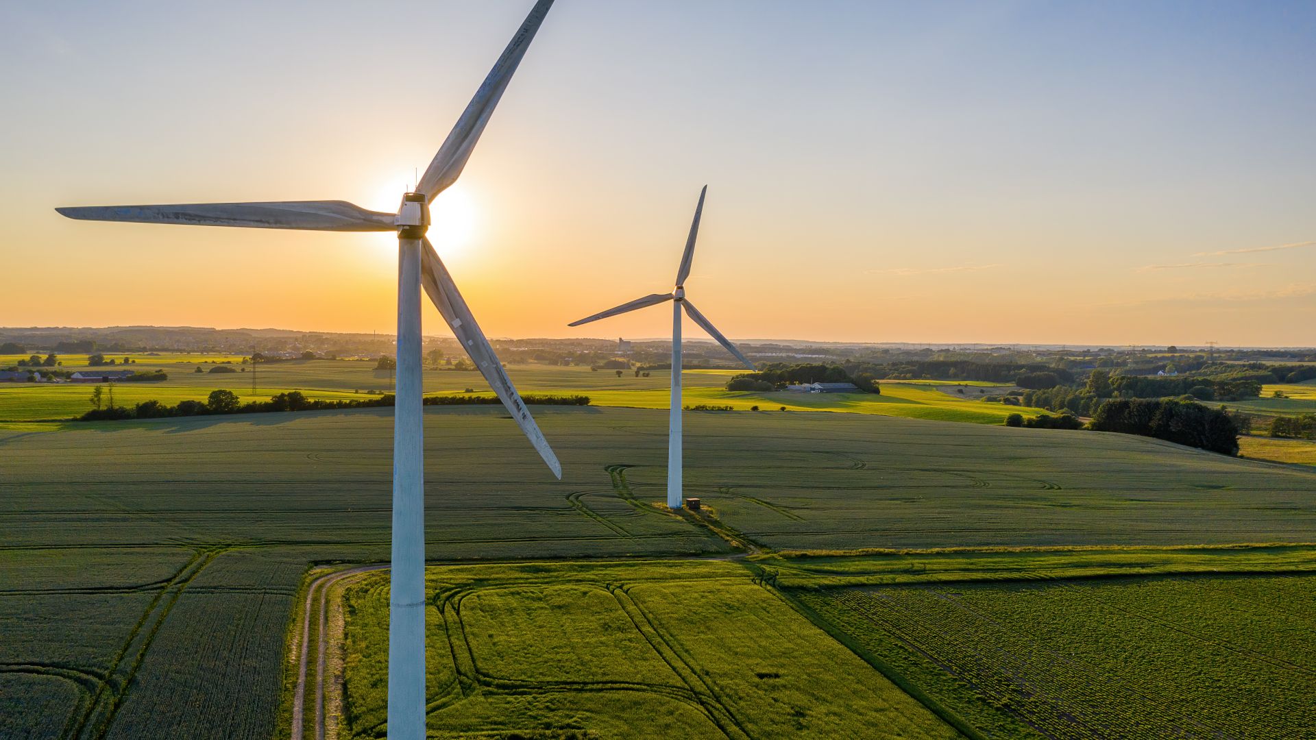 Two wind turbines standing in a vast green field at sunset, with the sun glowing behind one turbine and a rural landscape in the background.