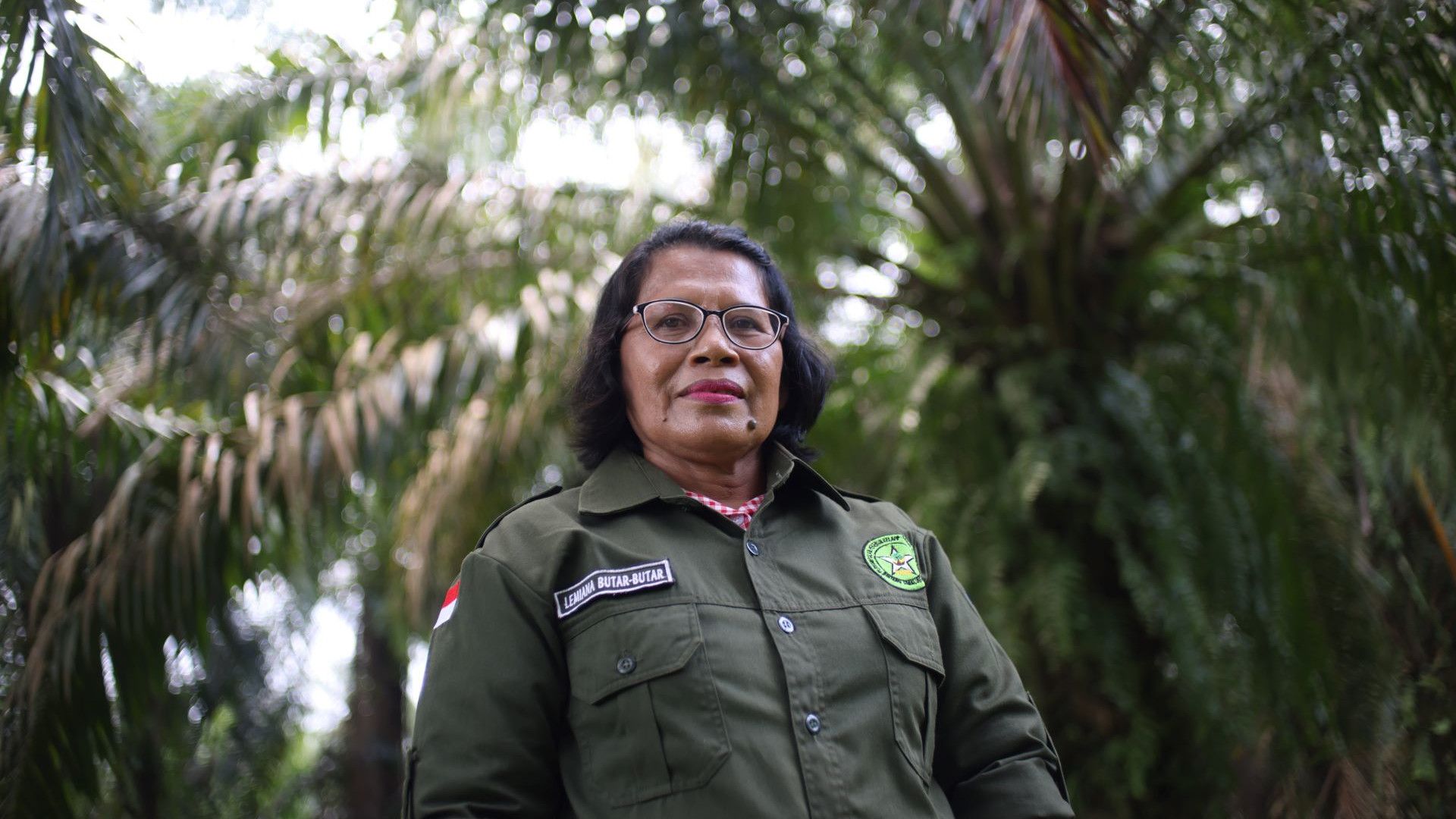 A woman standing in front on, looking at the camera wearing a green jacket. She is standing amongst palm oil trees.