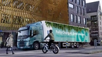 An electric truck with 'Fully Charged. Zero Emission. This truck is 100% electric.' written on its side, driving through a modern urban area with a cyclist and a pedestrian nearby, and contemporary buildings in the background.