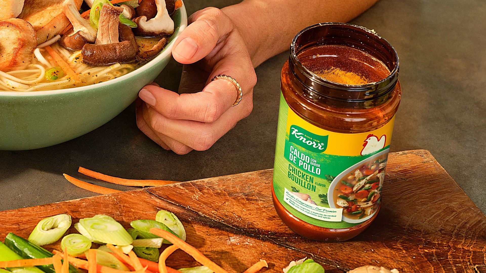 A jar of Knorr Chicken Bouillon on a wooden cutting board, next to a bowl of noodle broth.