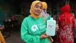Woman wearing long sleeved green t-shirt and yellow head scarf smiling at the camera holding a large bottle of green liquid product.