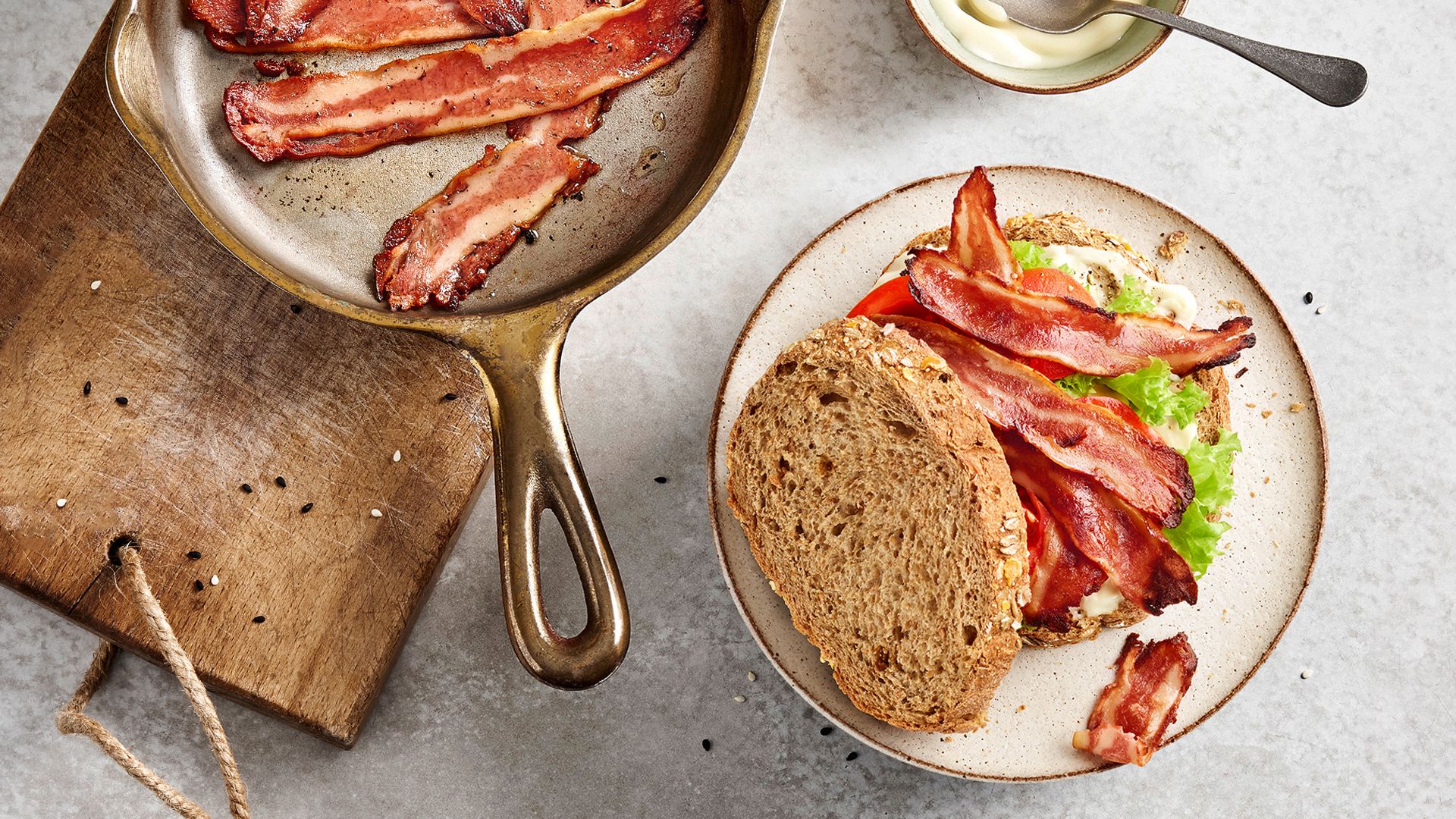Top view of sandwich placed on a plate with a pan of bacon on the side.