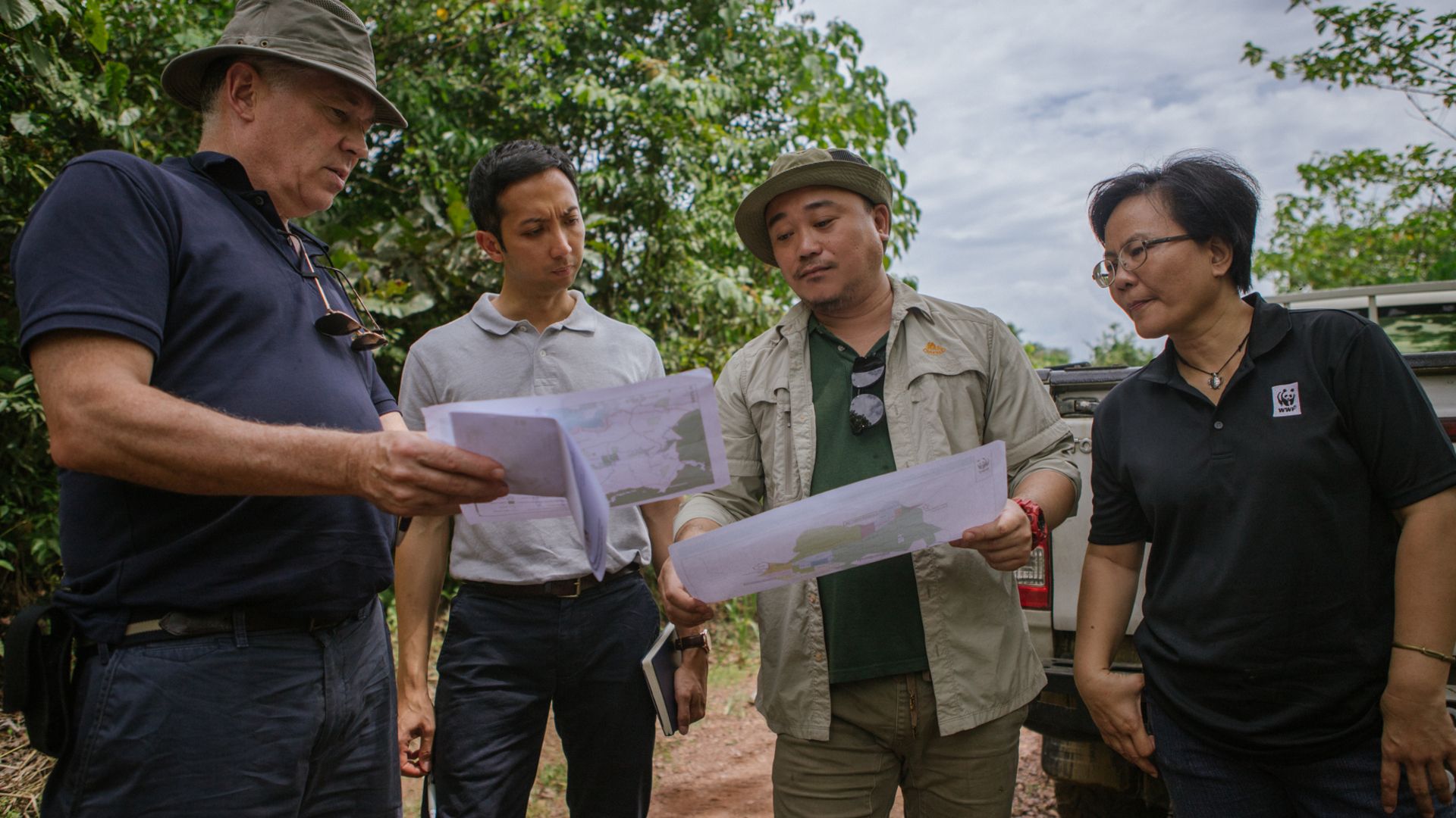Four individuals are engaged in a conversation and looking at printed maps in a field/plantation setting. The group includes people dressed in casual and outdoor work attire, one wearing a wide-brimmed hat and another in a shirt with a WWF logo.