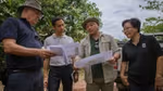 Four individuals are engaged in a conversation and looking at printed maps in a field/plantation setting. The group includes people dressed in casual and outdoor work attire, one wearing a wide-brimmed hat and another in a shirt with a WWF logo.