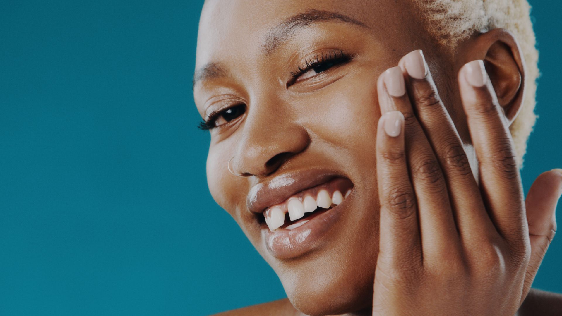 A woman with short, bleached hair applies skin cream to her cheek. She smiles at the camera.