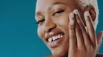 A woman with short, bleached hair applies skin cream to her cheek. She smiles at the camera.