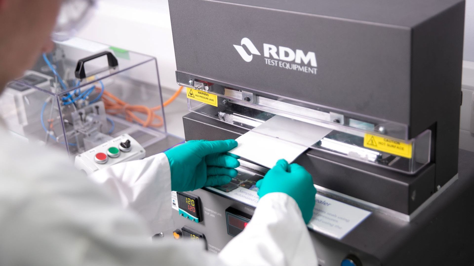 A packaging lab technician wearing green gloves places a sample sheet into an RDM Test Equipment machine. The technician is holding the sample with two hands. The background shows additional lab apparatus and tools.
