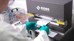 A packaging lab technician wearing green gloves places a sample sheet into an RDM Test Equipment machine. The technician is holding the sample with two hands. The background shows additional lab apparatus and tools.