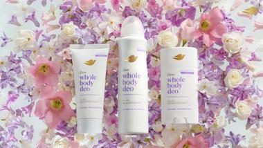 Three Dove Whole Body Deo products arranged on a bed of pastel flowers in shades of pink, purple, and white.