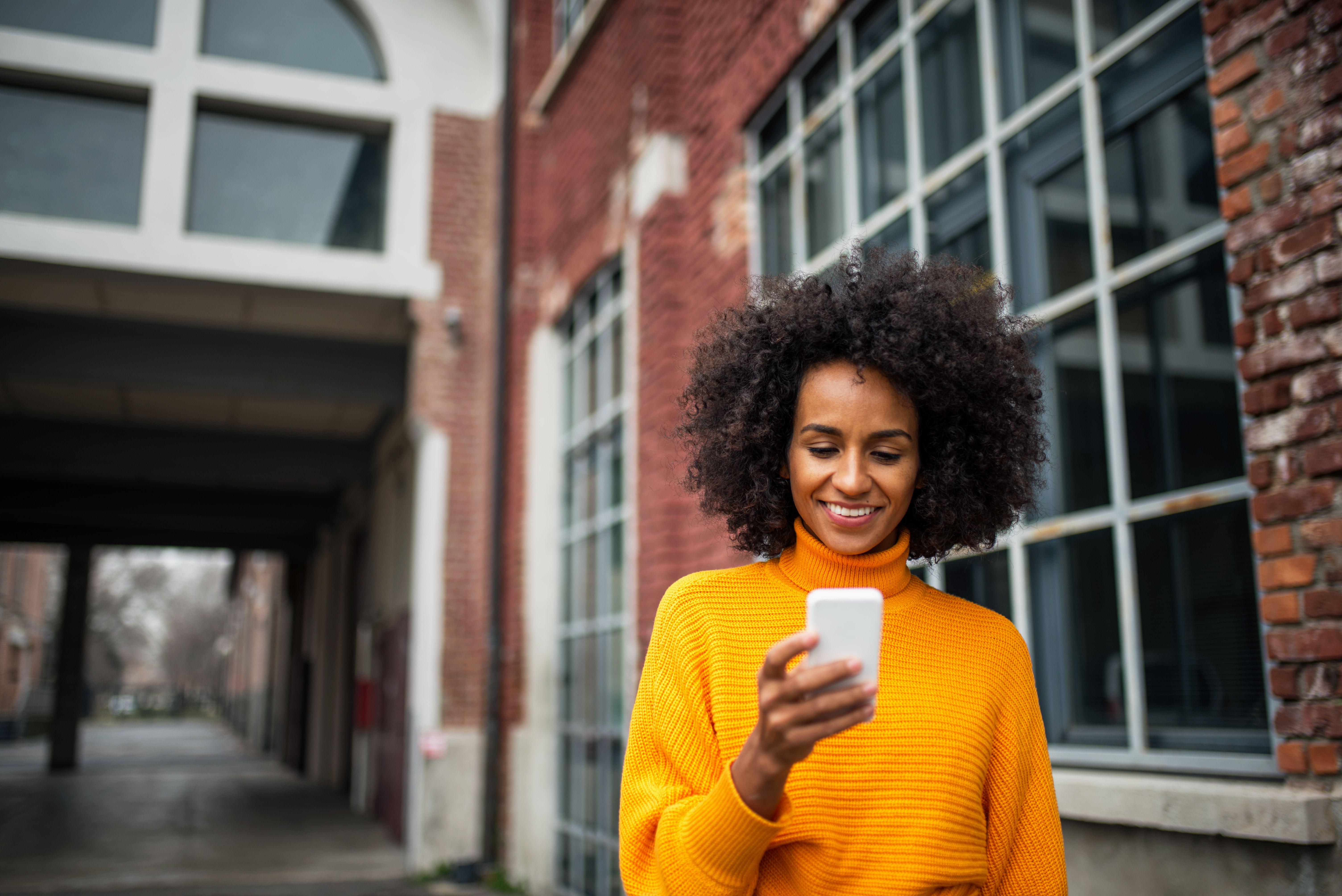 A woman in a city smiles as she looks down at her phone