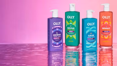Four colourful Olly Mood + Skin body wash bottles stand on a reflective surface against a pink gradient background.