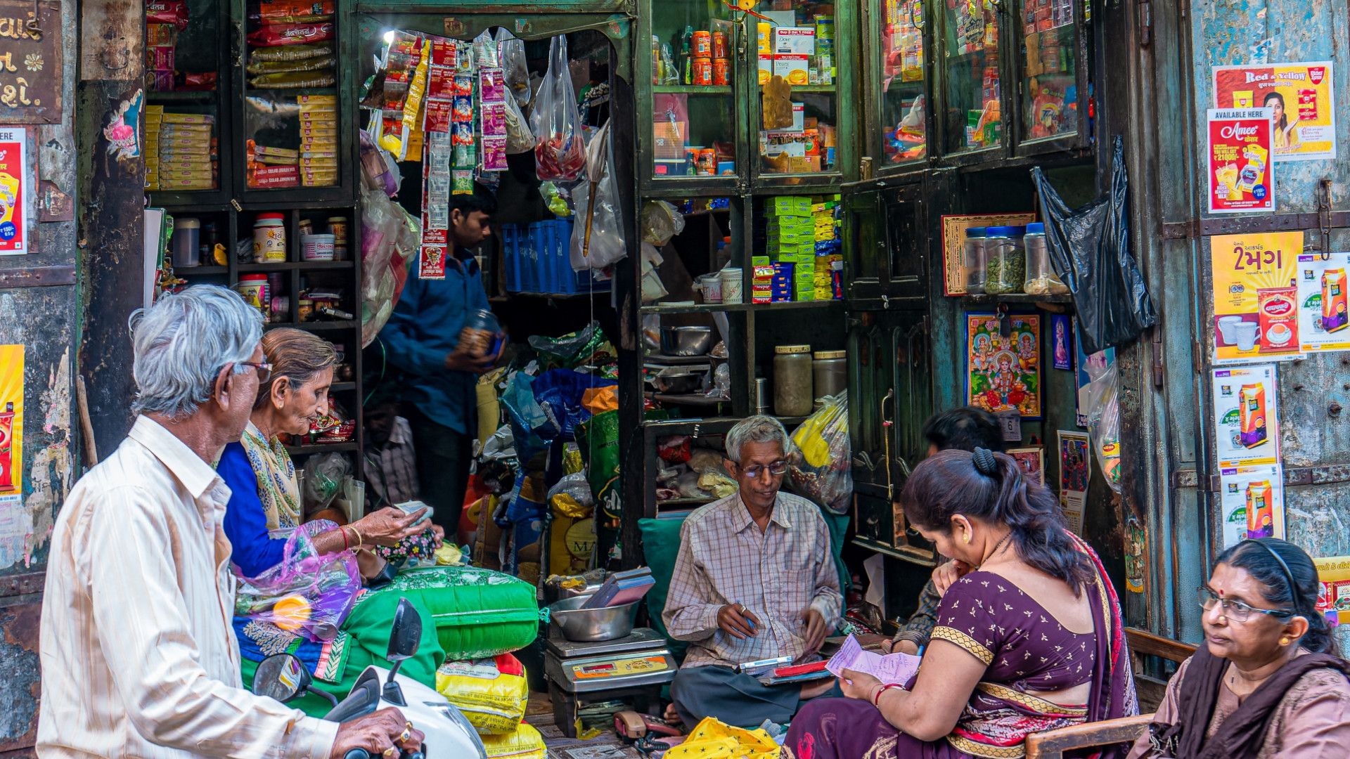 Three men and three women in saris sit outside a small store in rural India, surrounded by colourful products.