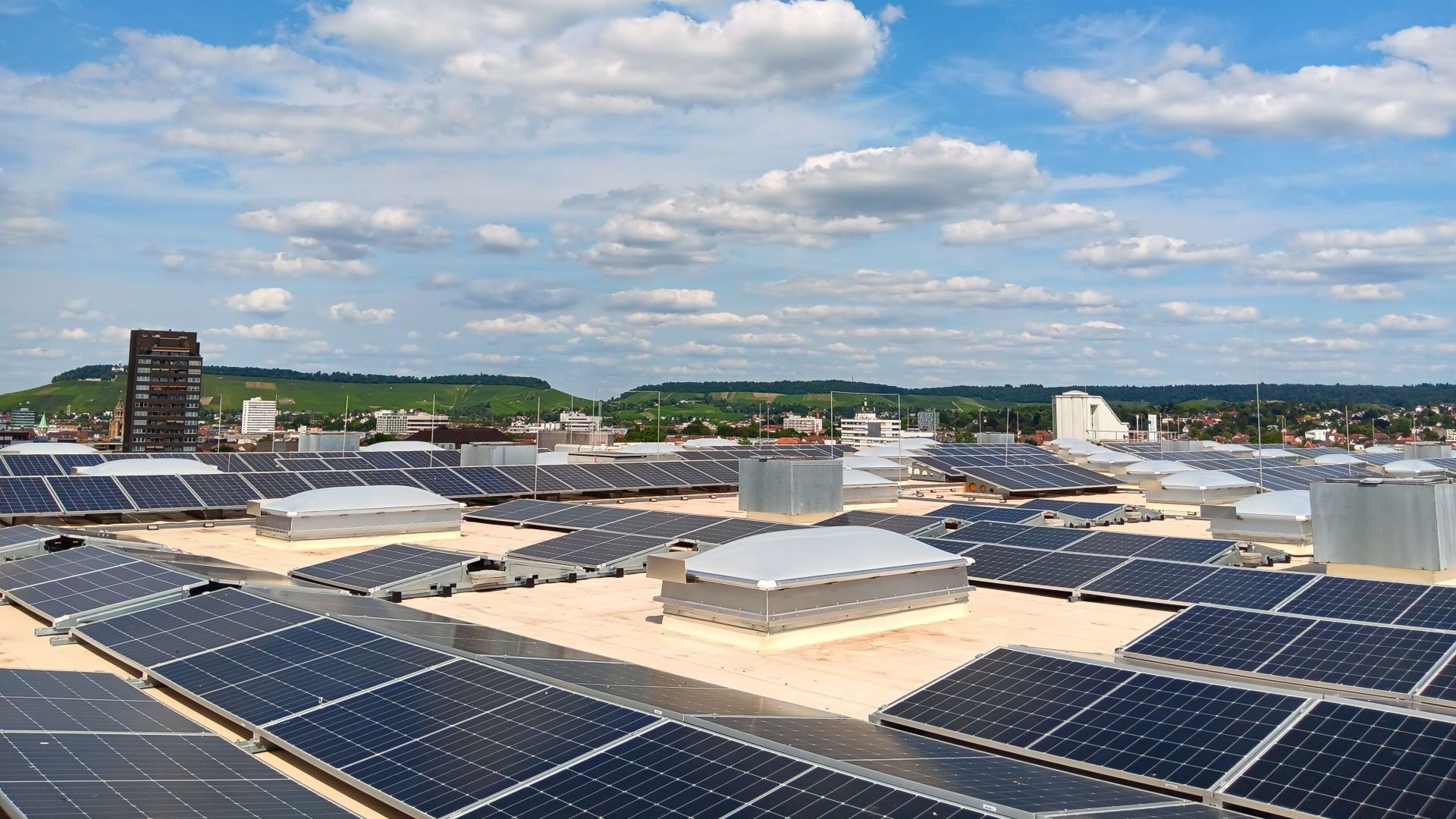 Solar panels cover a large rooftop overlooking a city and green hills.