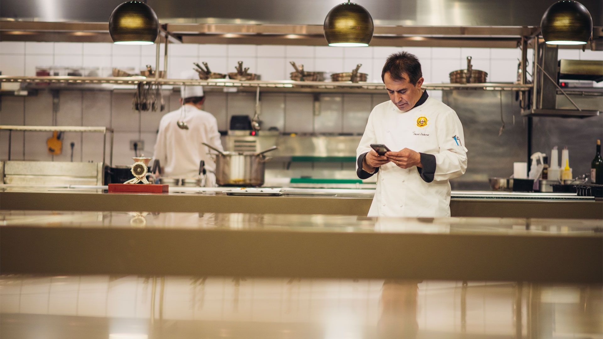 A chef consulting his phone in a professional kitchen while another chef works  in the background 