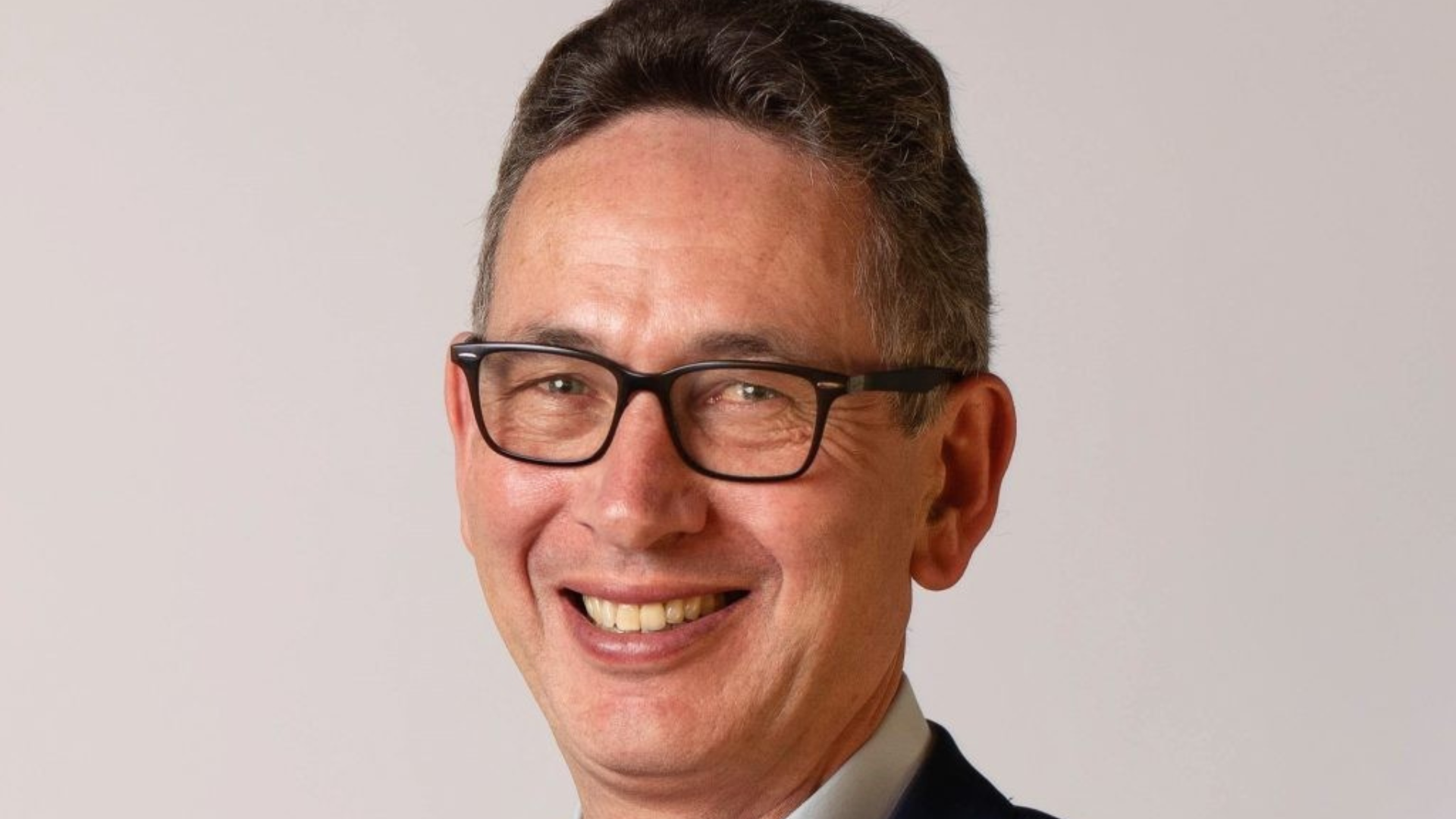 A headshot of Marcus Gover. Marcus has short dark hair and blue eyes, and is wearing a light blue open collar shirt and navy blue jacket. He is smiling at the camera.